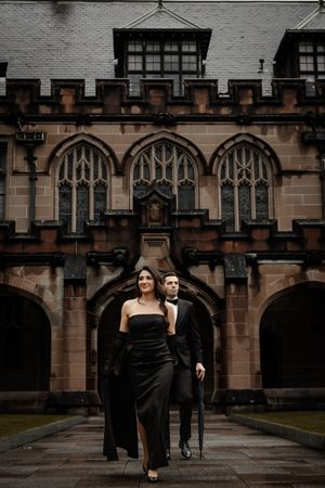 Couple in black formal attire walking through Gothic stone archway during engagement shoot.