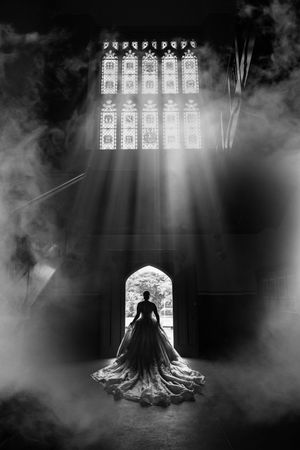 Dramatic black and white silhouette of bride in ballgown framed by light rays and mist in gothic church.