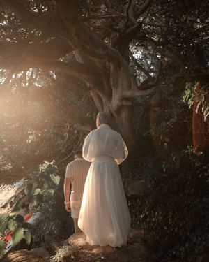 Couple stands beneath a sunlit ancient tree in a lush garden, bride wearing a flowing white skirt.
