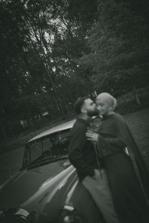 Black and white artistic photo of two grooms sharing a kiss beside a vintage car in a forest.