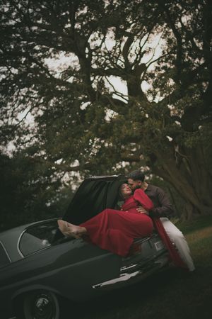 Woman in red dress reclines on a vintage car hood as her partner leans in beneath a large tree.