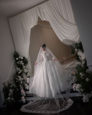 Bride in a stunning ball gown wedding dress with lace veil stands beneath a draped white arch with floral arrangements.