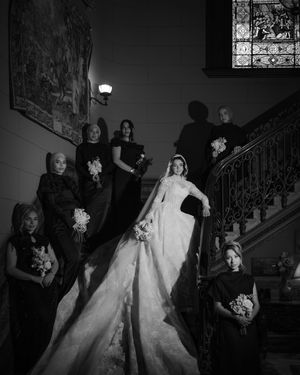 Black and white photo of bride in lace gown with bridesmaids in black dresses on an elegant staircase.