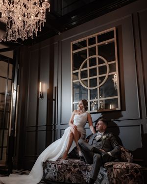 Bride and groom relax on ornate sofa beneath a crystal chandelier in a dramatic dark-walled luxury venue.