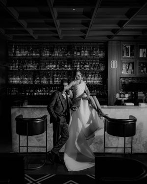 Black and white photo of bride and groom posing at a sophisticated bar with bottle-lined shelves.