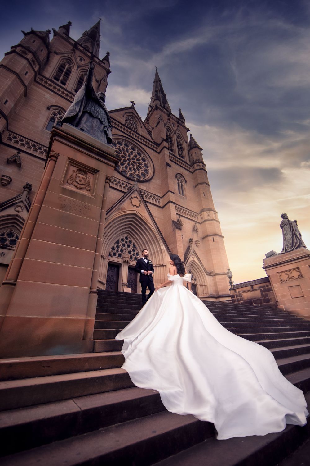 Bride and groom on cathedral steps at sunset, wedding dress train dramatically flowing in the breeze.