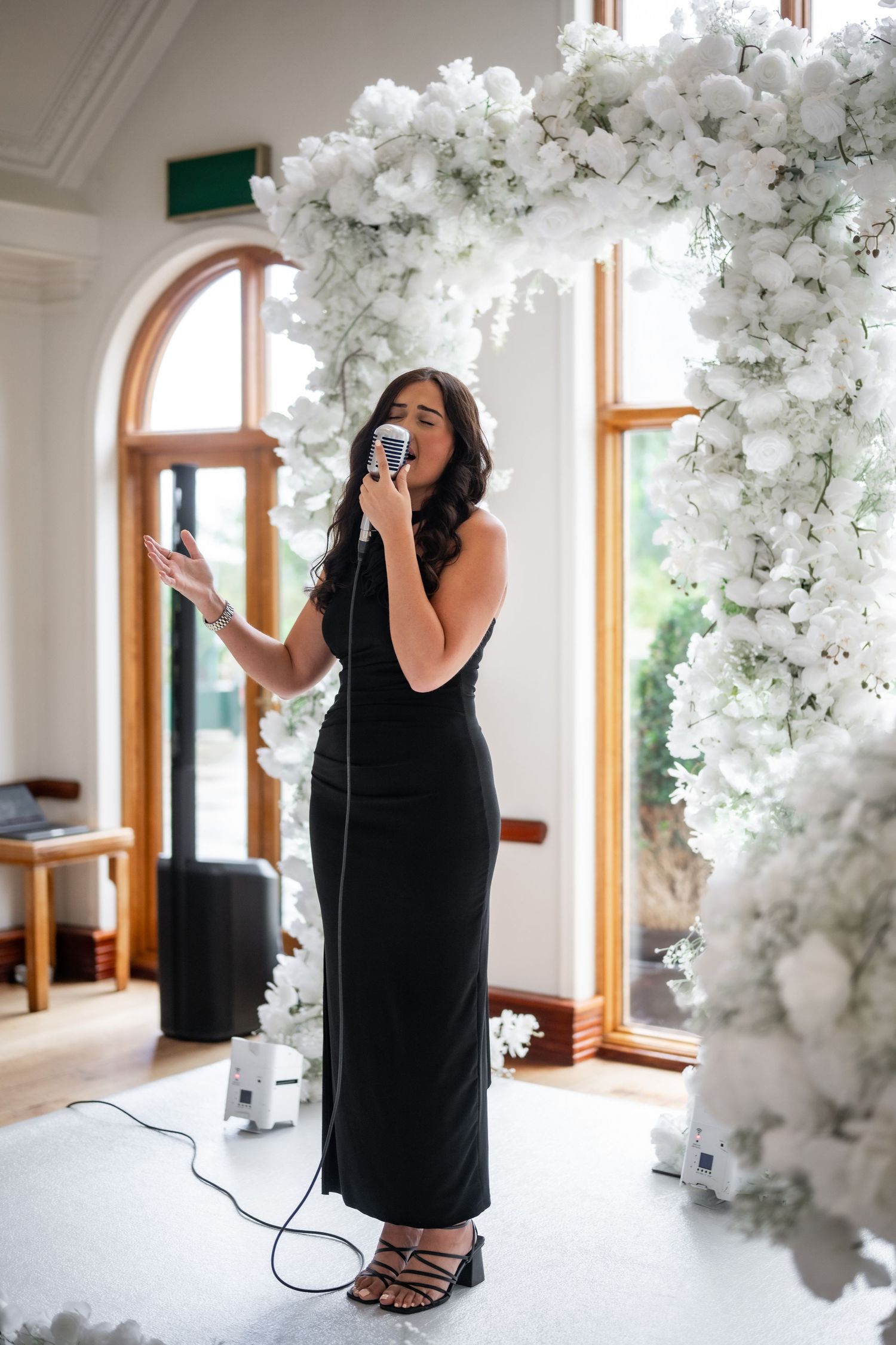 A singer in a long black dress performs under an elegant white floral arch by large arched windows. Chelsea Johnson