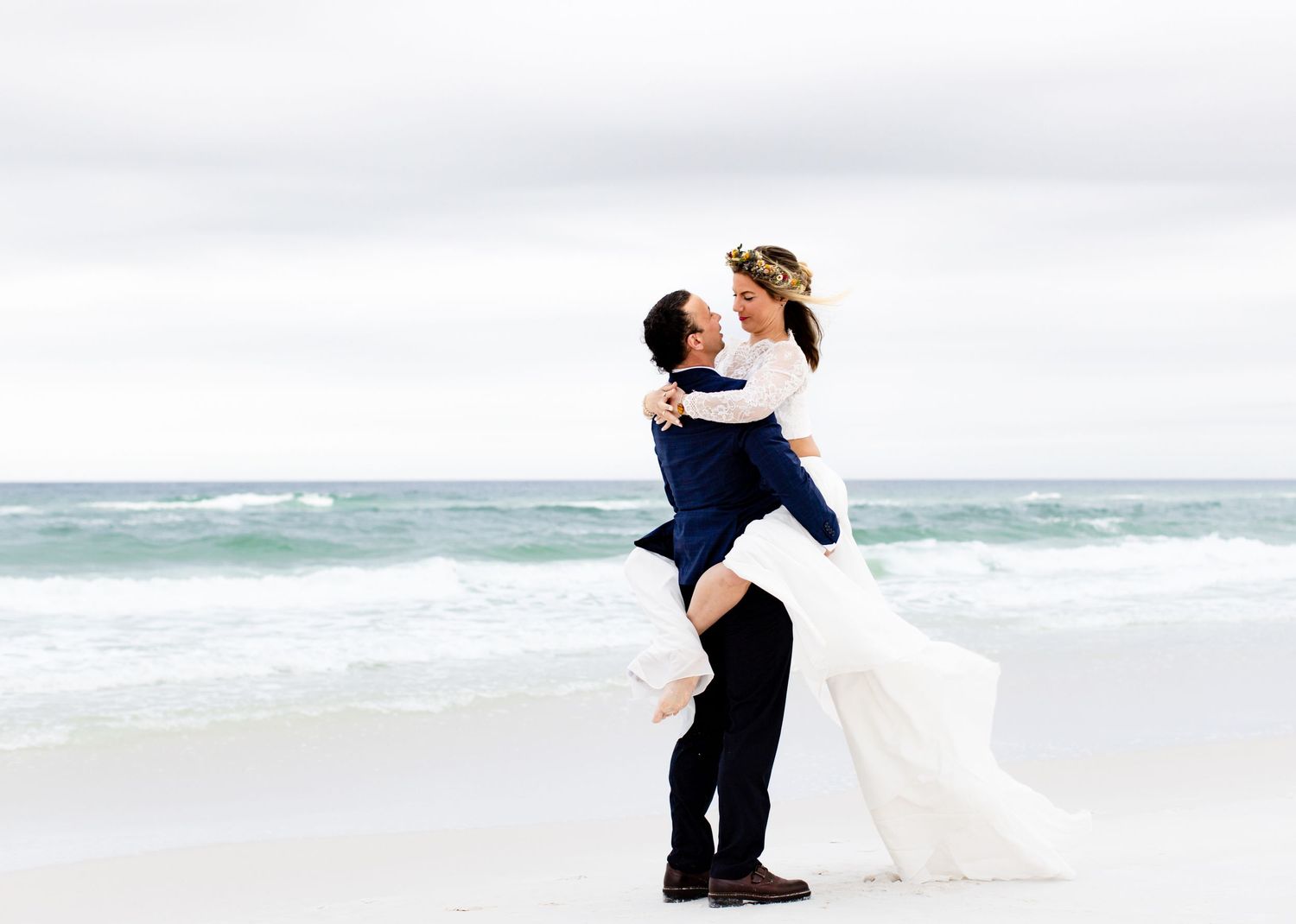 A groom lifts his bride in a flowing white dress on a beach.