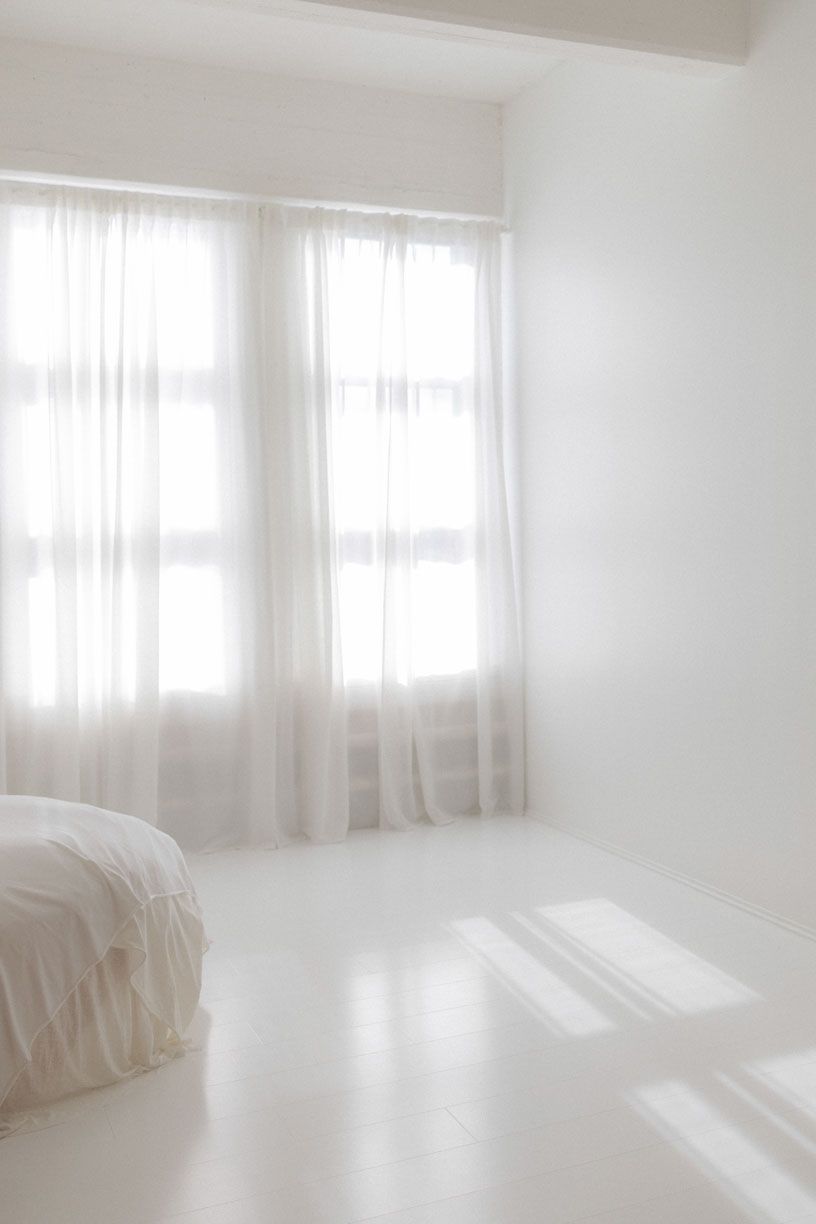 Minimalist white bedroom interior with sheer curtains filtering soft natural light and creating peaceful ambiance.