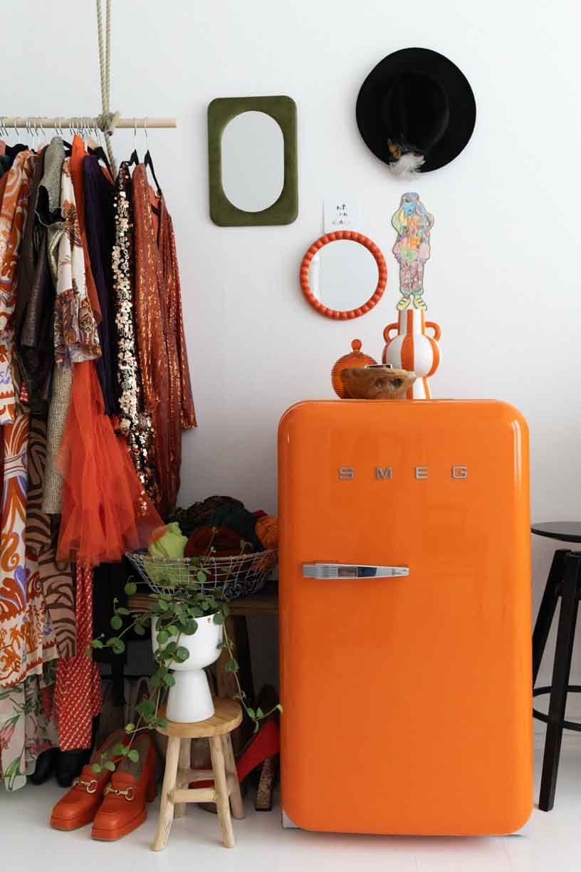 Retro orange SMEG refrigerator styled with wooden stool and clothing rack in a minimalist interior setting.