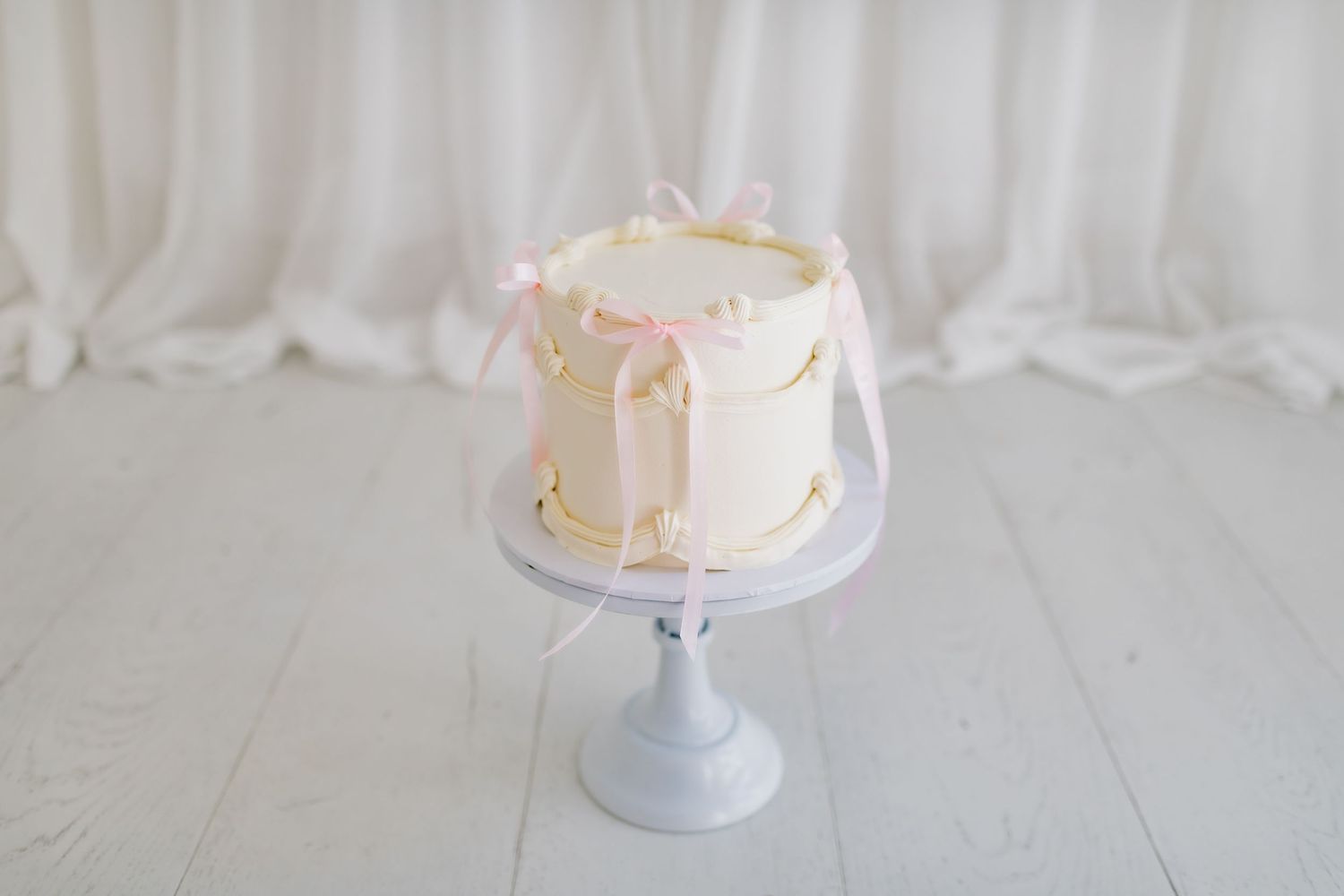 A simple white cake with pink drips and decorative elements sits atop a pale blue cake stand.