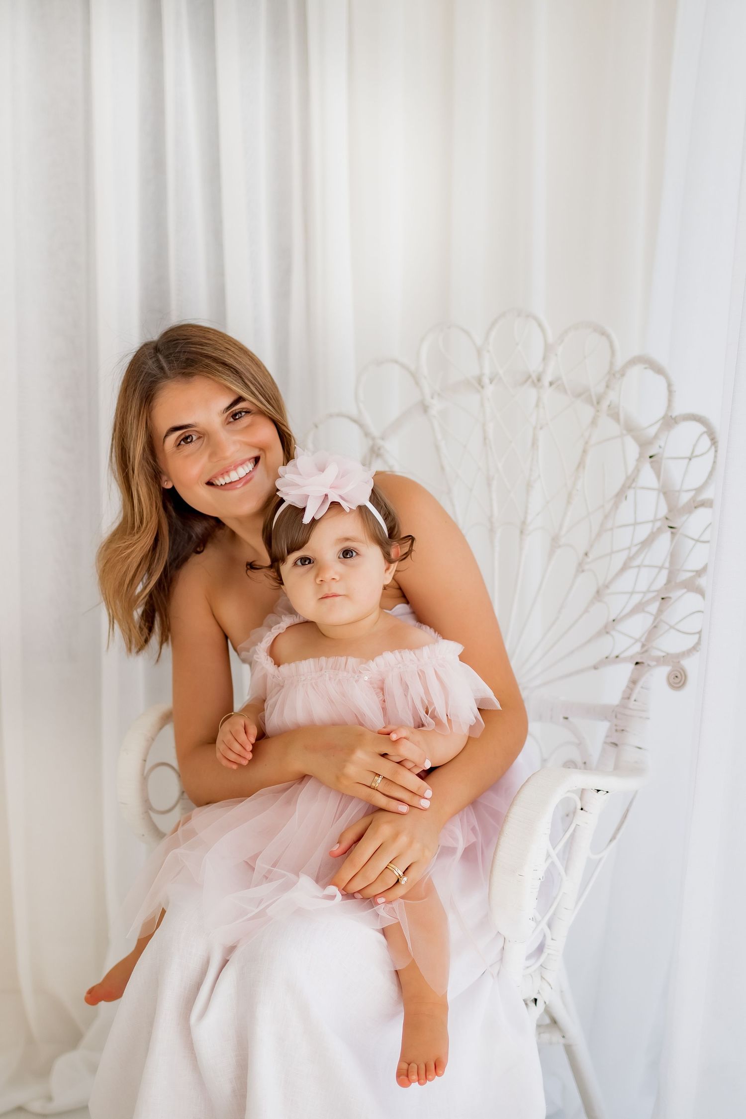 Two people in pink and white outfits sitting in a decorative white peacock chair against white curtains.
