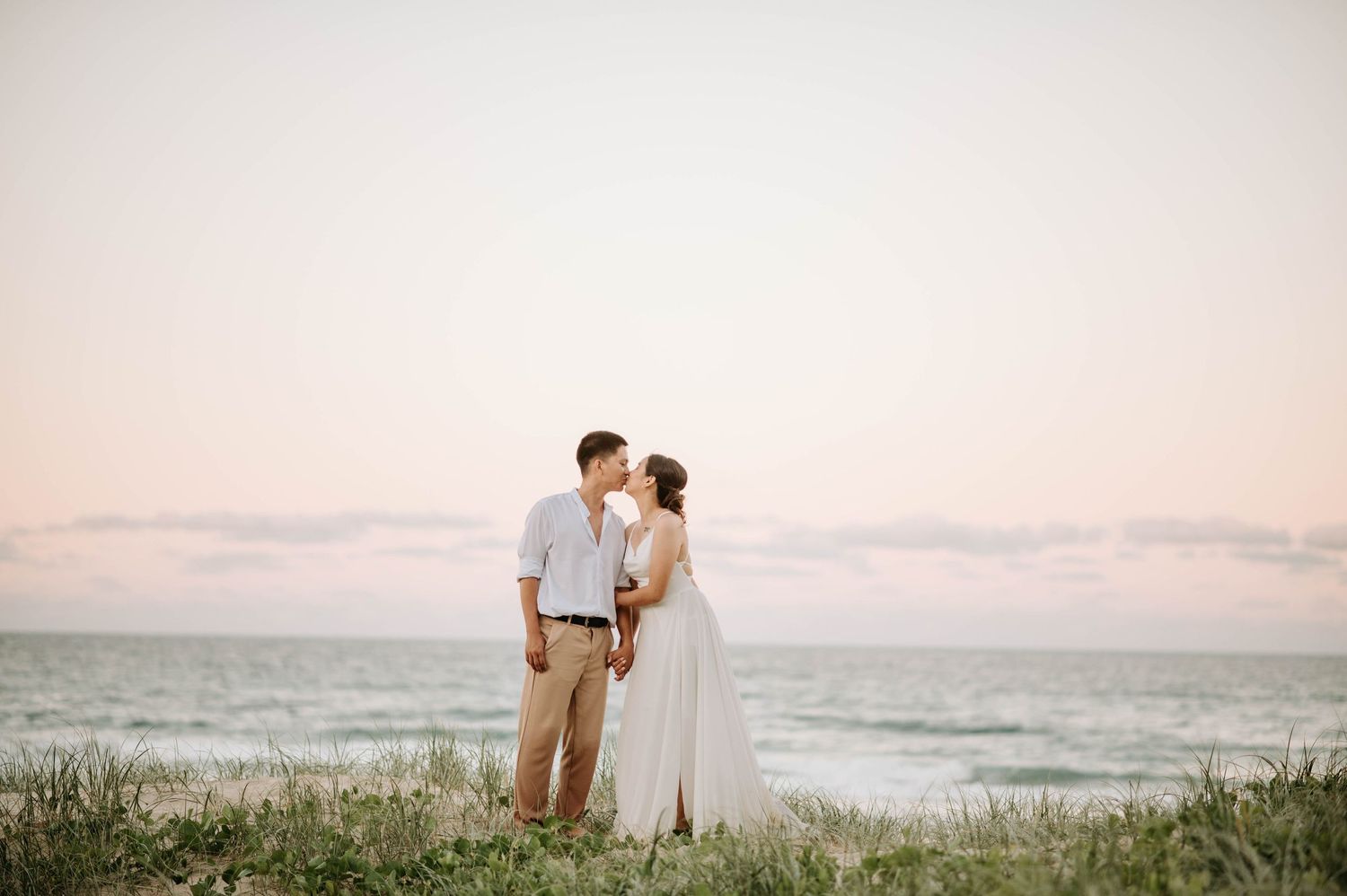 A romantic beach engagement photo at sunset shows a couple standing together on grassy dunes by the ocean.