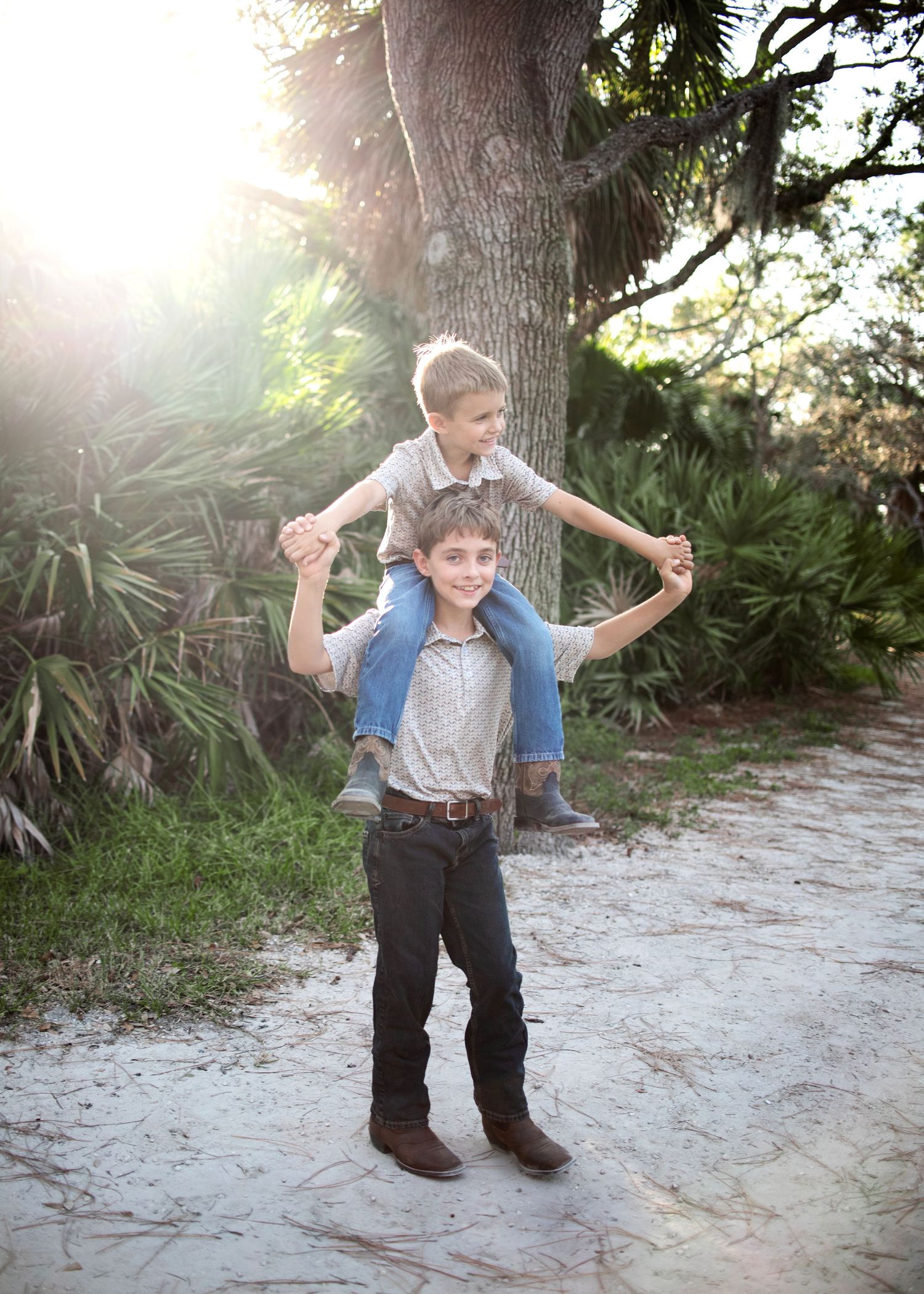 Older boy carries younger boy on shoulders on a sandy path near tropical trees.