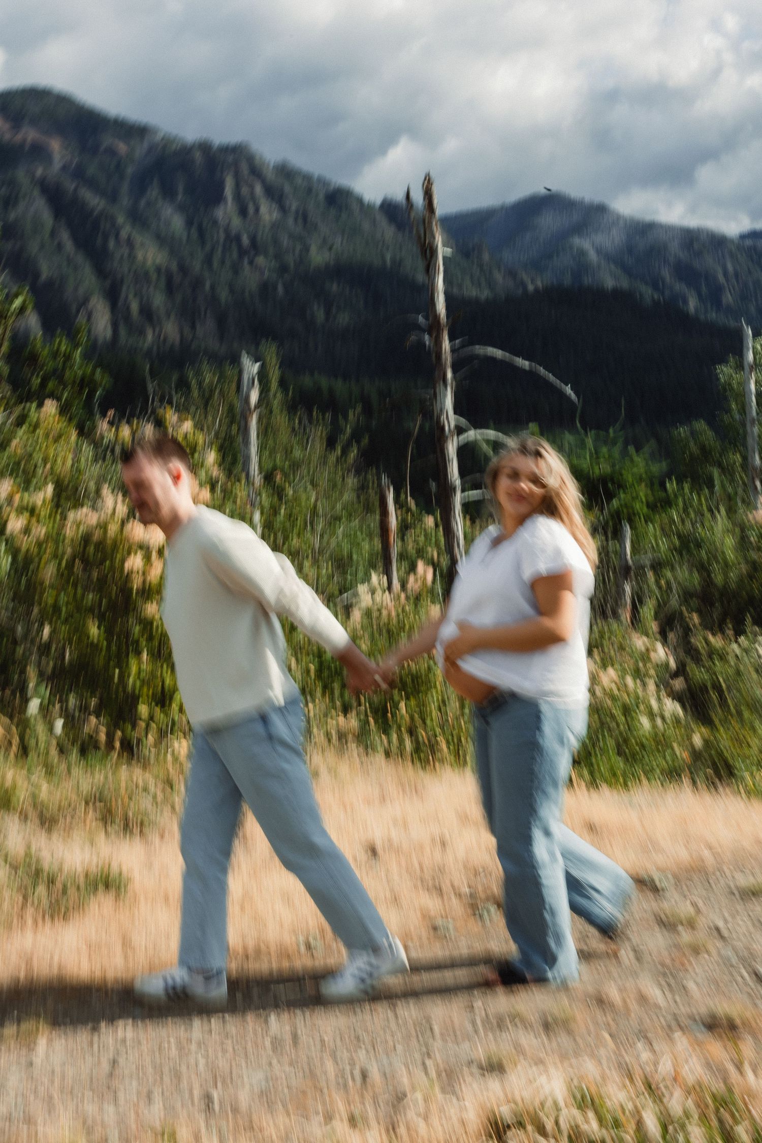 Two people playfully dance and spin together in a field with dramatic mountain peaks and cloudy skies in the background.