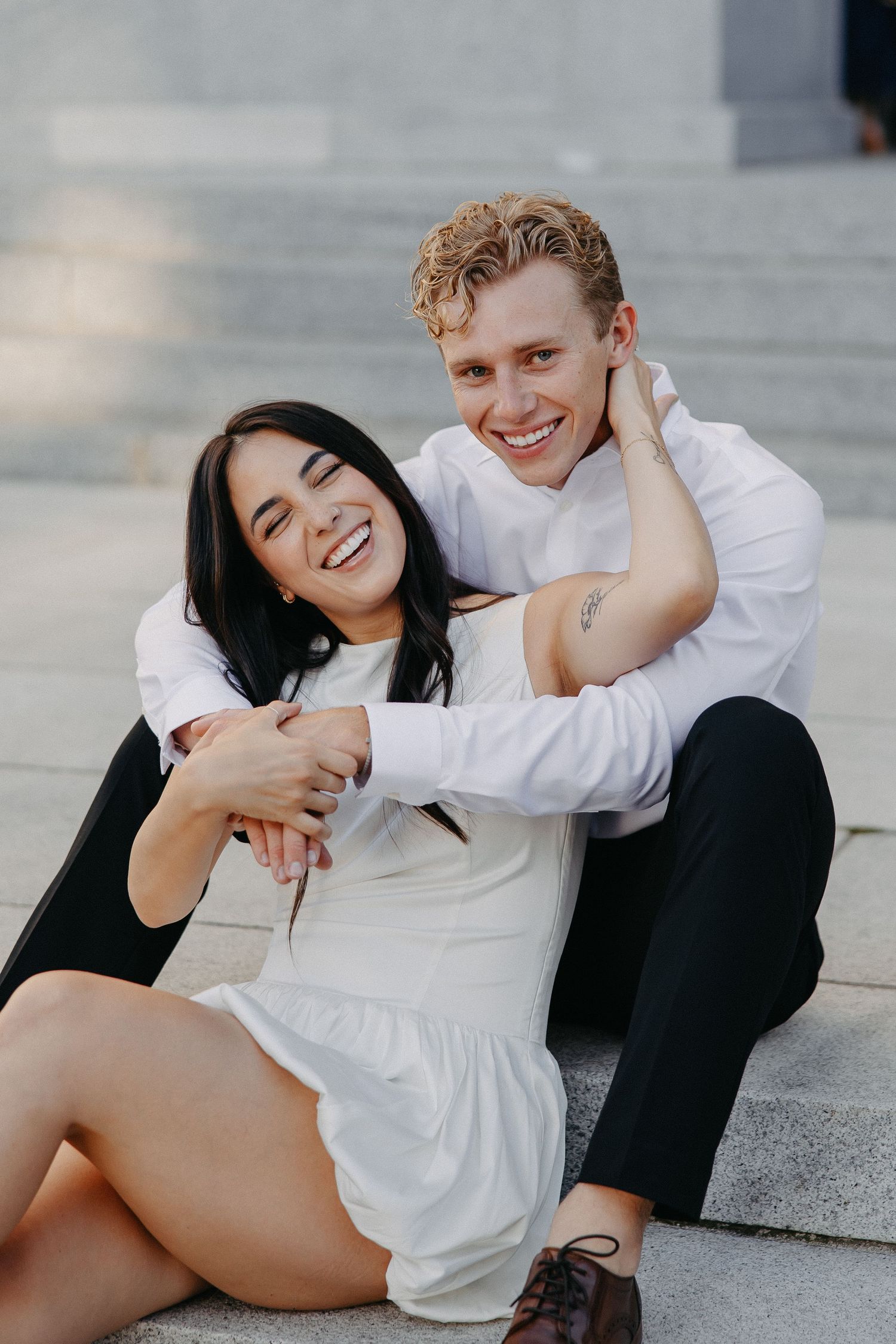 A couple in white and black outfits share playful candid moments while sitting on concrete steps.