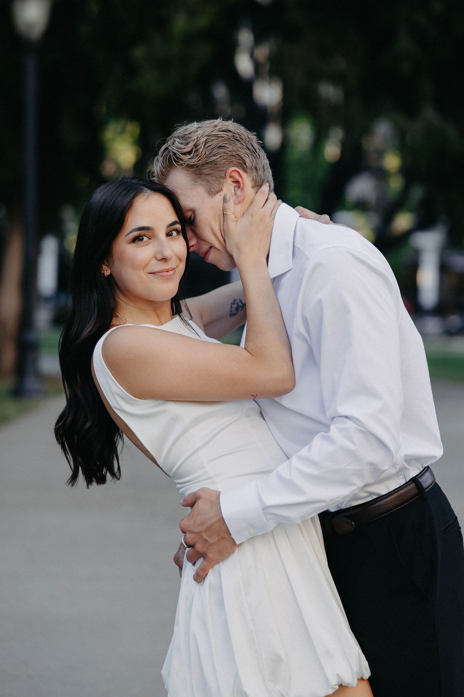 A romantic couple in white clothing embrace and share an intimate moment outdoors in a park setting.
