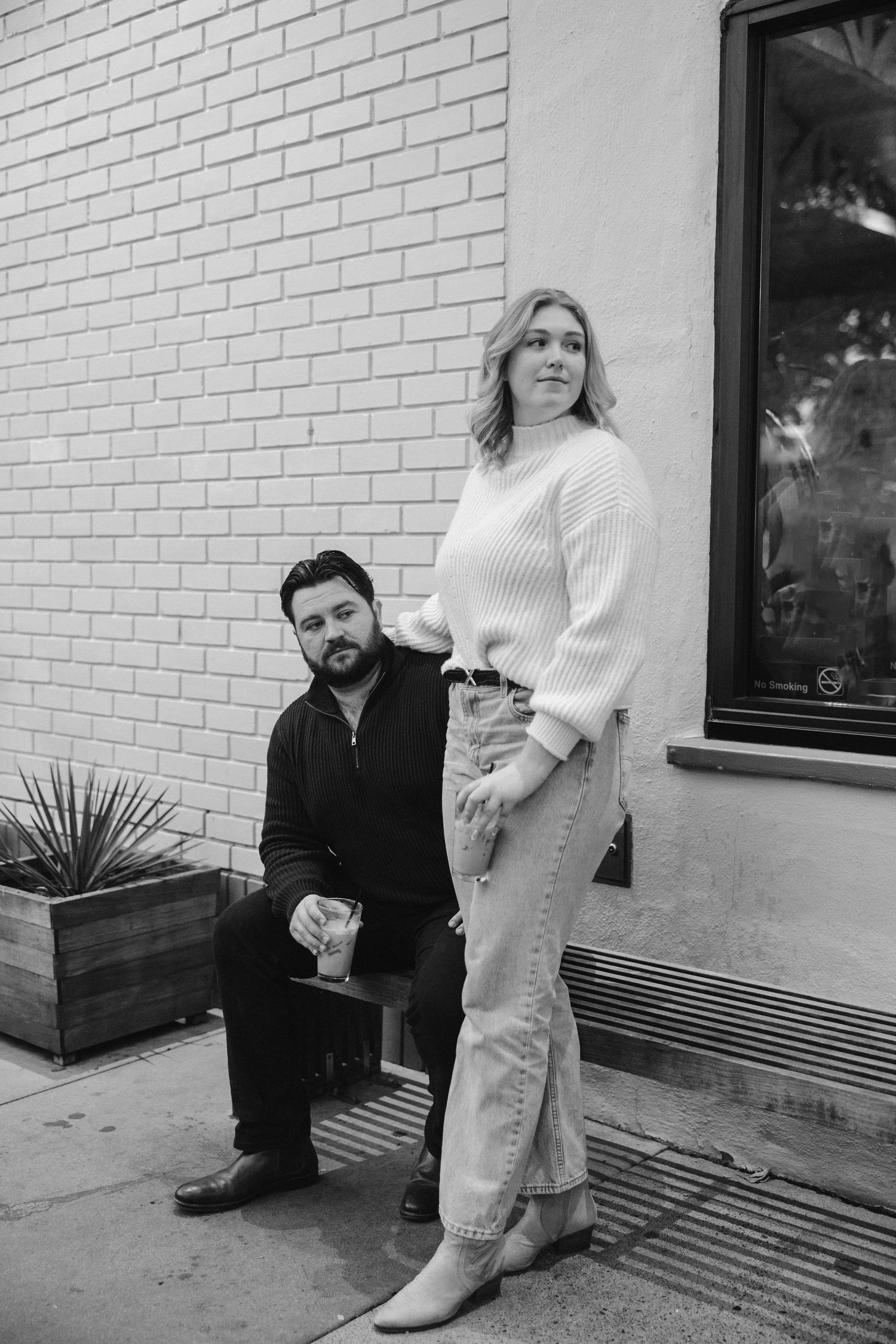A couple poses casually against a white brick wall in casual winter attire.
