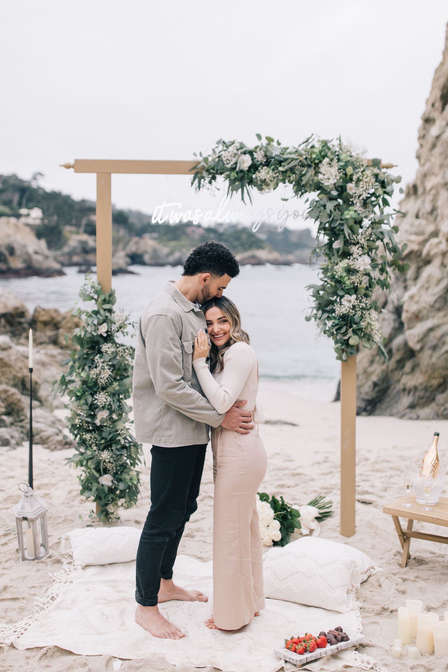 Intimate barefoot couple embracing beneath wooden arch adorned with white flowers on rocky beach.