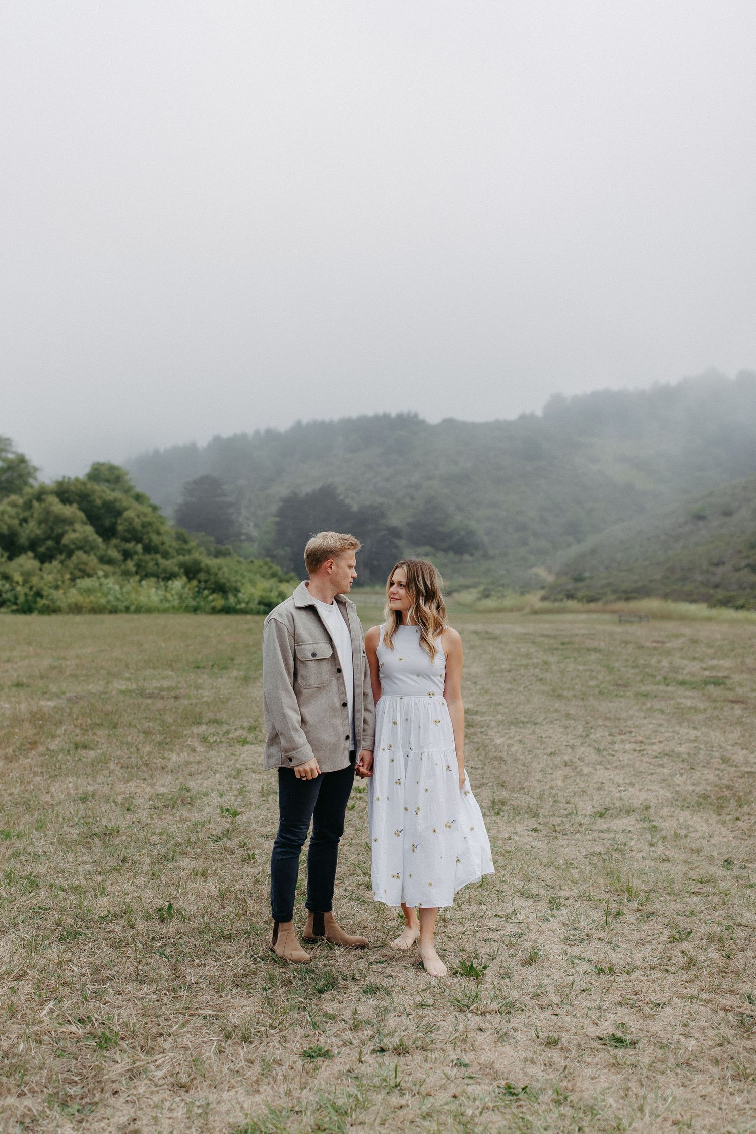 A couple stands together in an open field with misty mountains behind them.