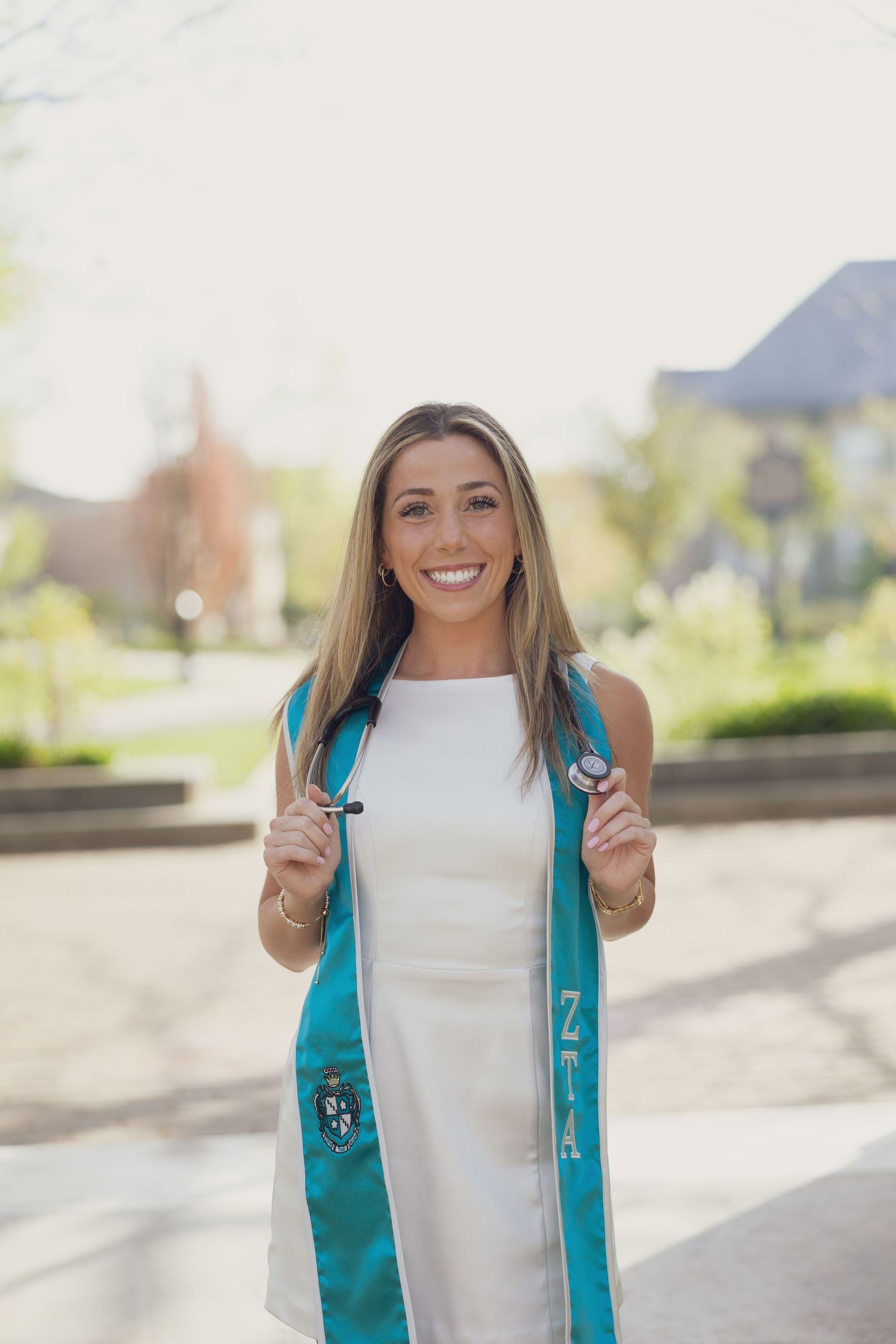 Graduate wearing a white dress and teal graduation stole stands outdoors smiling with campus buildings in background.