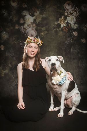 Young woman in black dress and floral crown poses with white and brown dog wearing flower collar against dark floral backdrop.