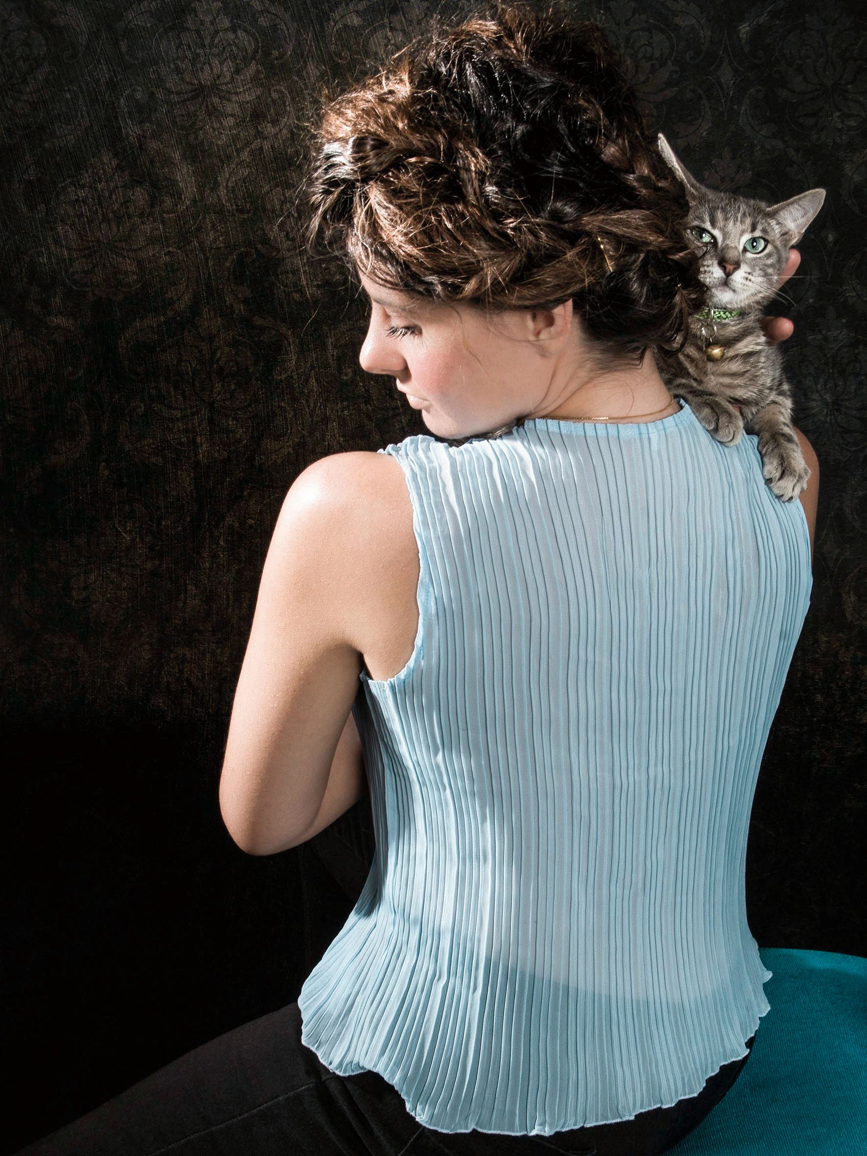Person in light blue pleated top holding a gray cat against dark background.