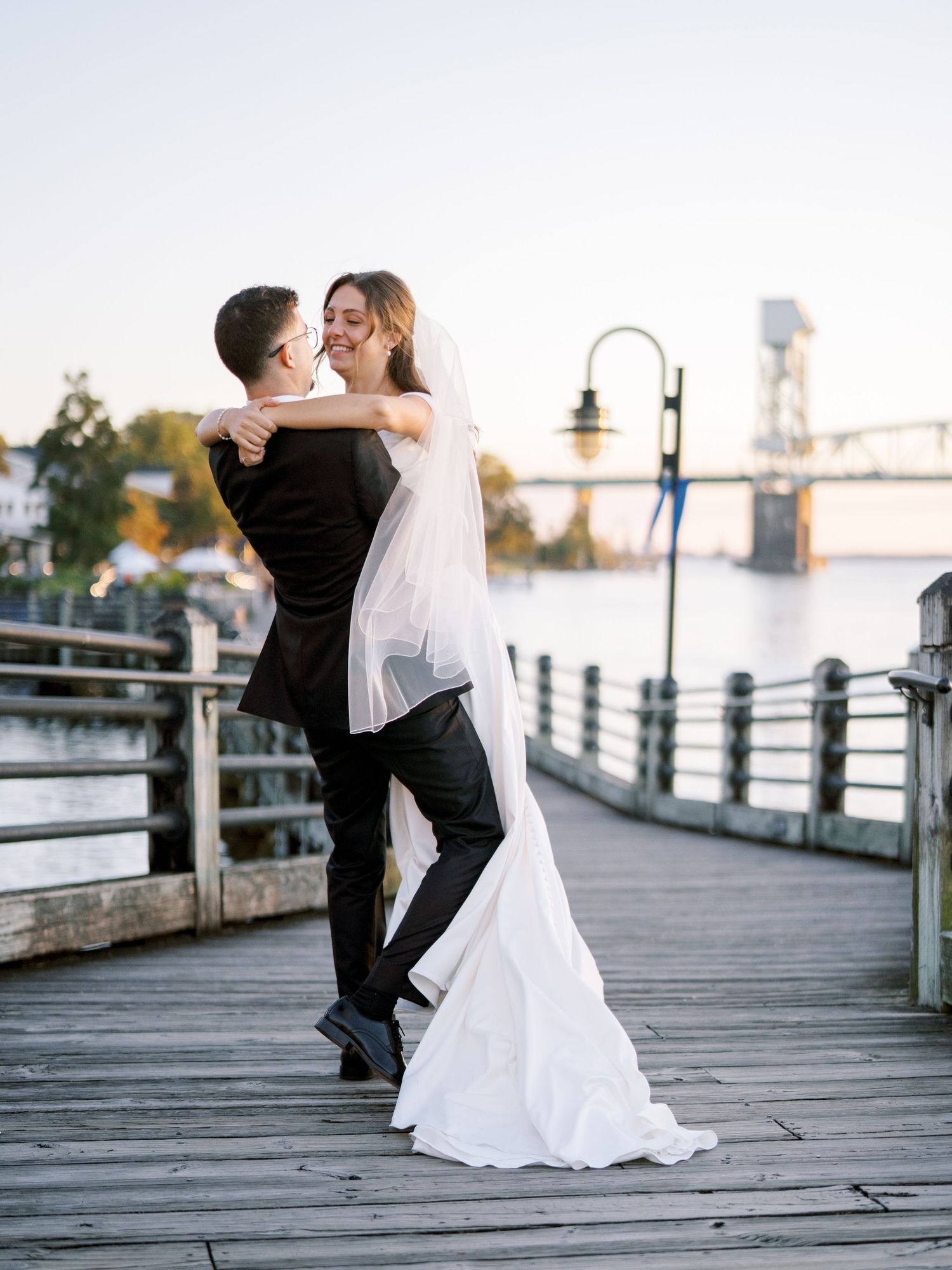 Romantic embrace on a waterfront boardwalk with industrial bridge in background.