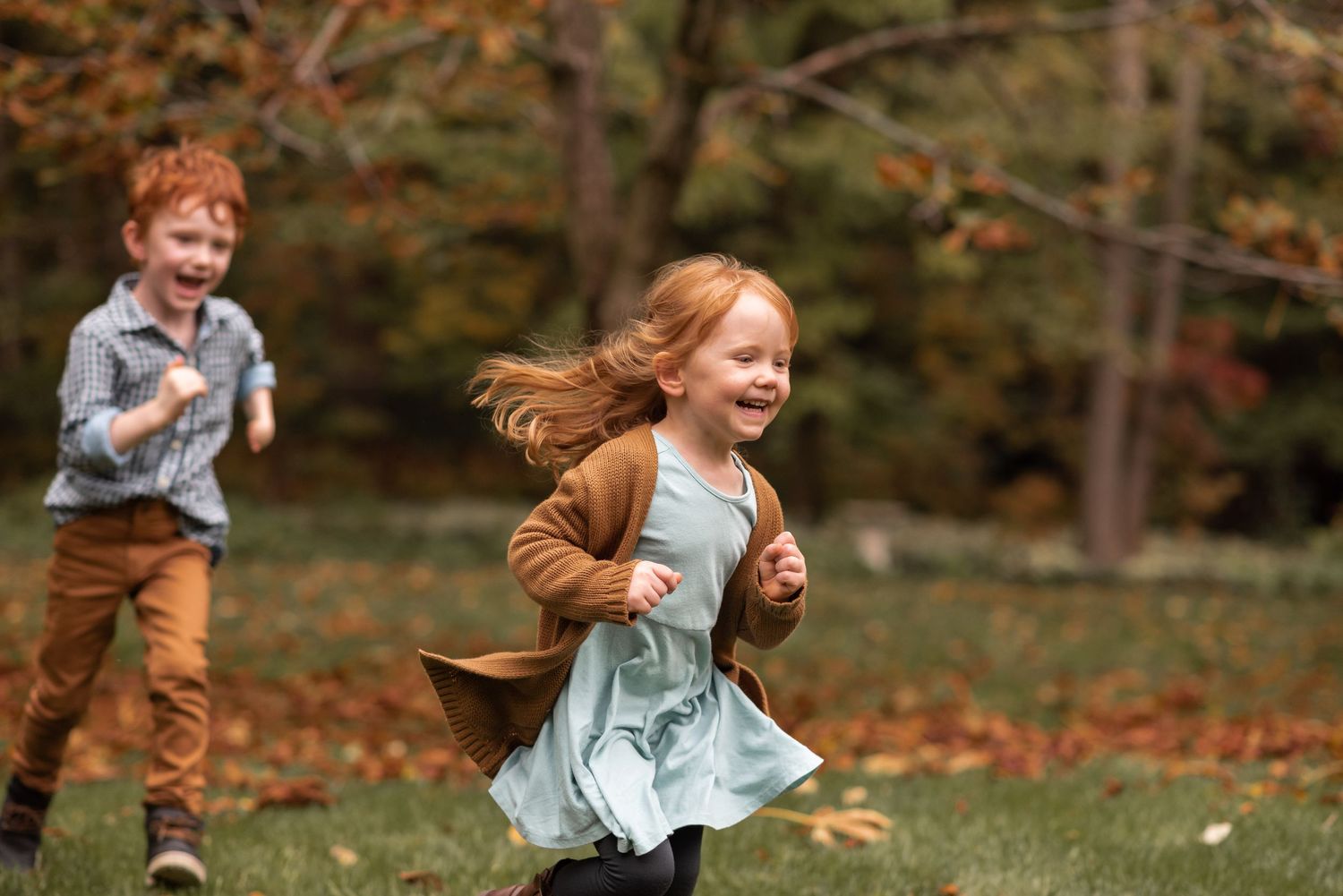 Children running and playing in autumn leaves on a grassy lawn surrounded by fall foliage.