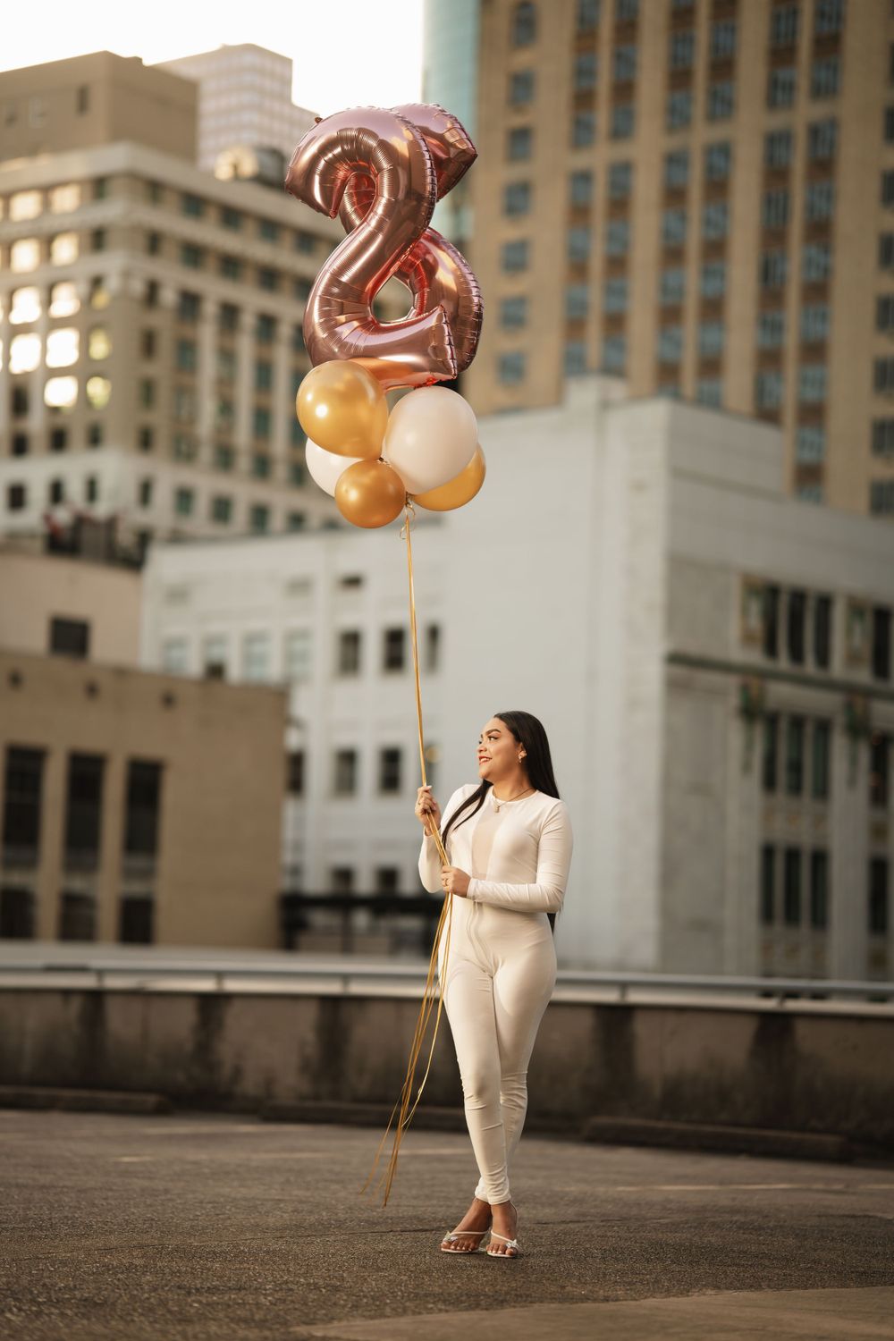 Celebratory rooftop scene with rose gold number balloons and cream outfit against urban cityscape backdrop.