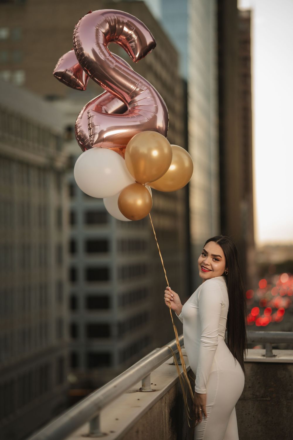 Elegant woman in white dress holds rose gold number 2 balloon with gold and white balloon bouquet on urban rooftop.