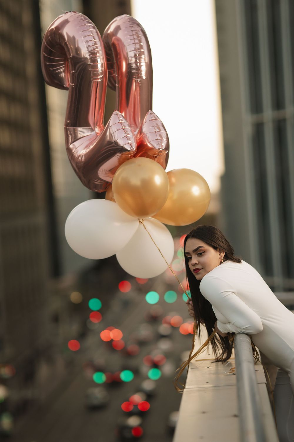 Person in white sweater holding rose gold, gold and white balloons on city street with blurred lights in background.