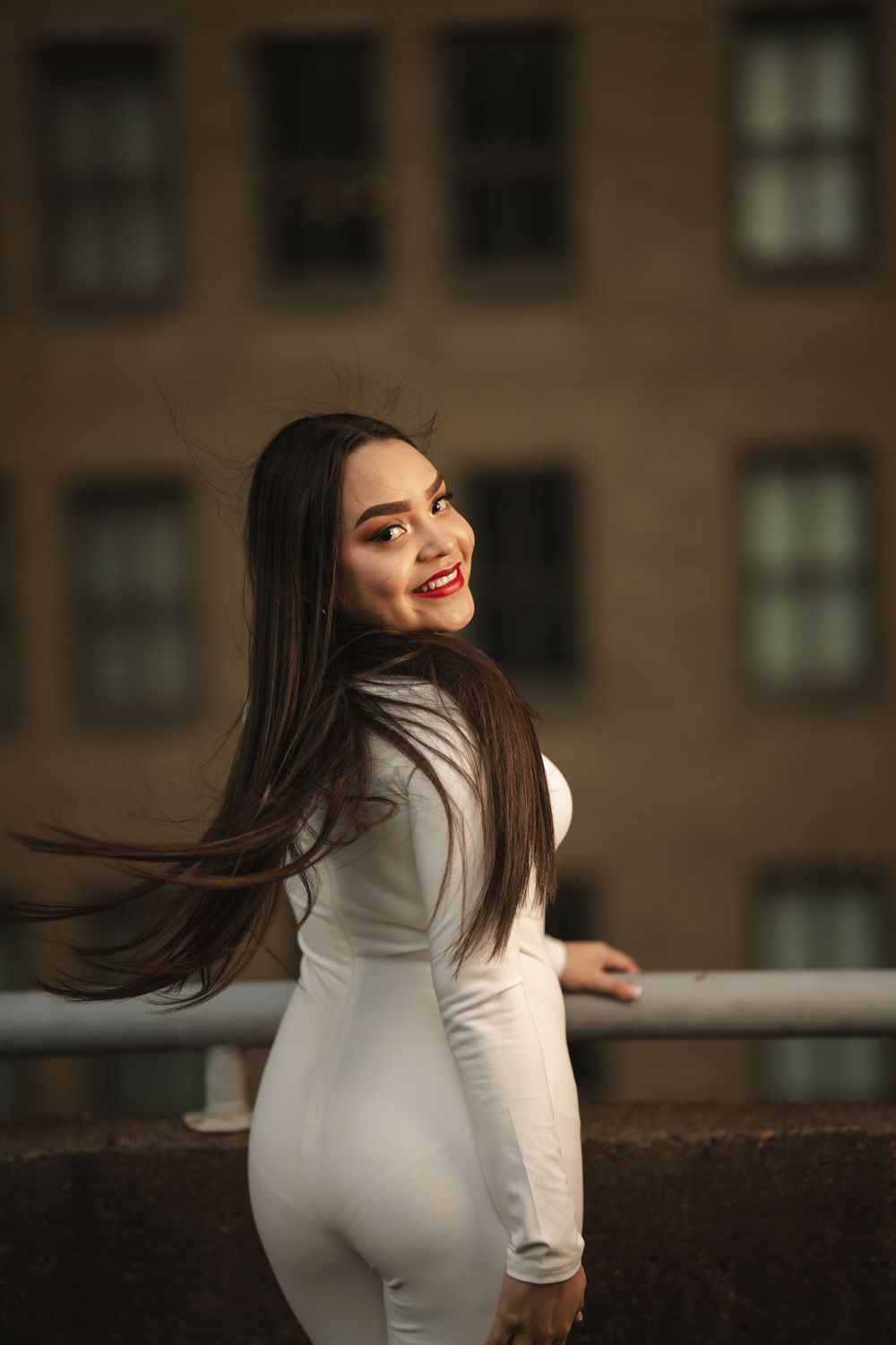 A person in a fitted white dress smiles while posing outdoors against an urban backdrop with flowing dark hair.
