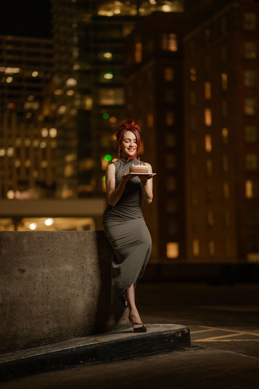 A woman in a fitted gray dress holds a tablet device while posing against a cityscape backdrop at night.