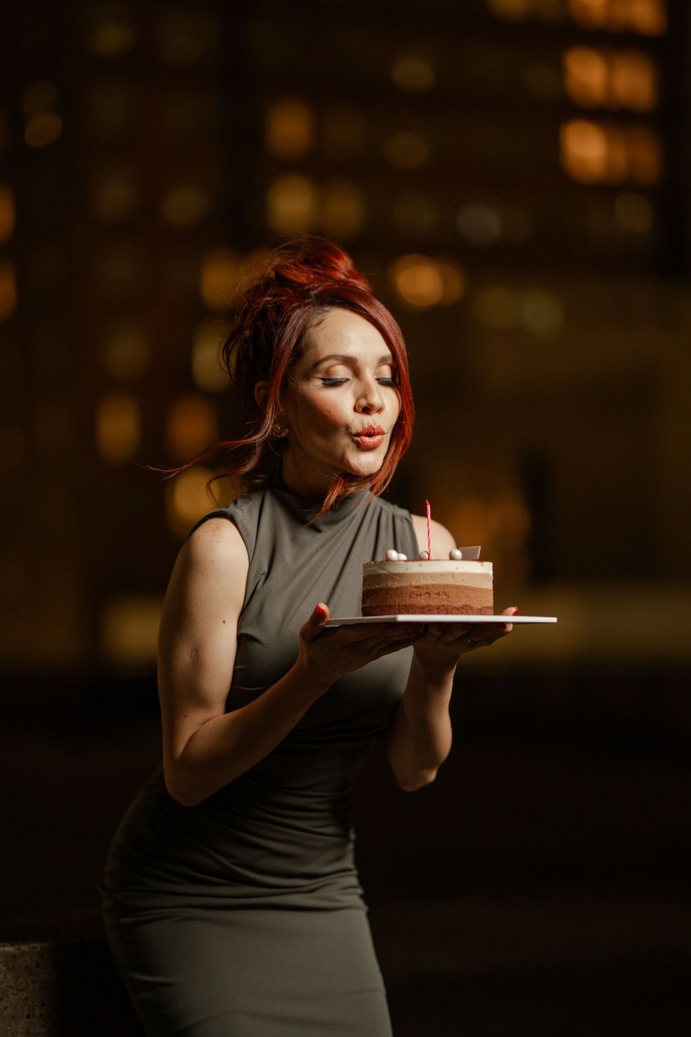 Woman in gray dress holds chocolate cake with candle while smiling in warm evening lighting against bokeh background.