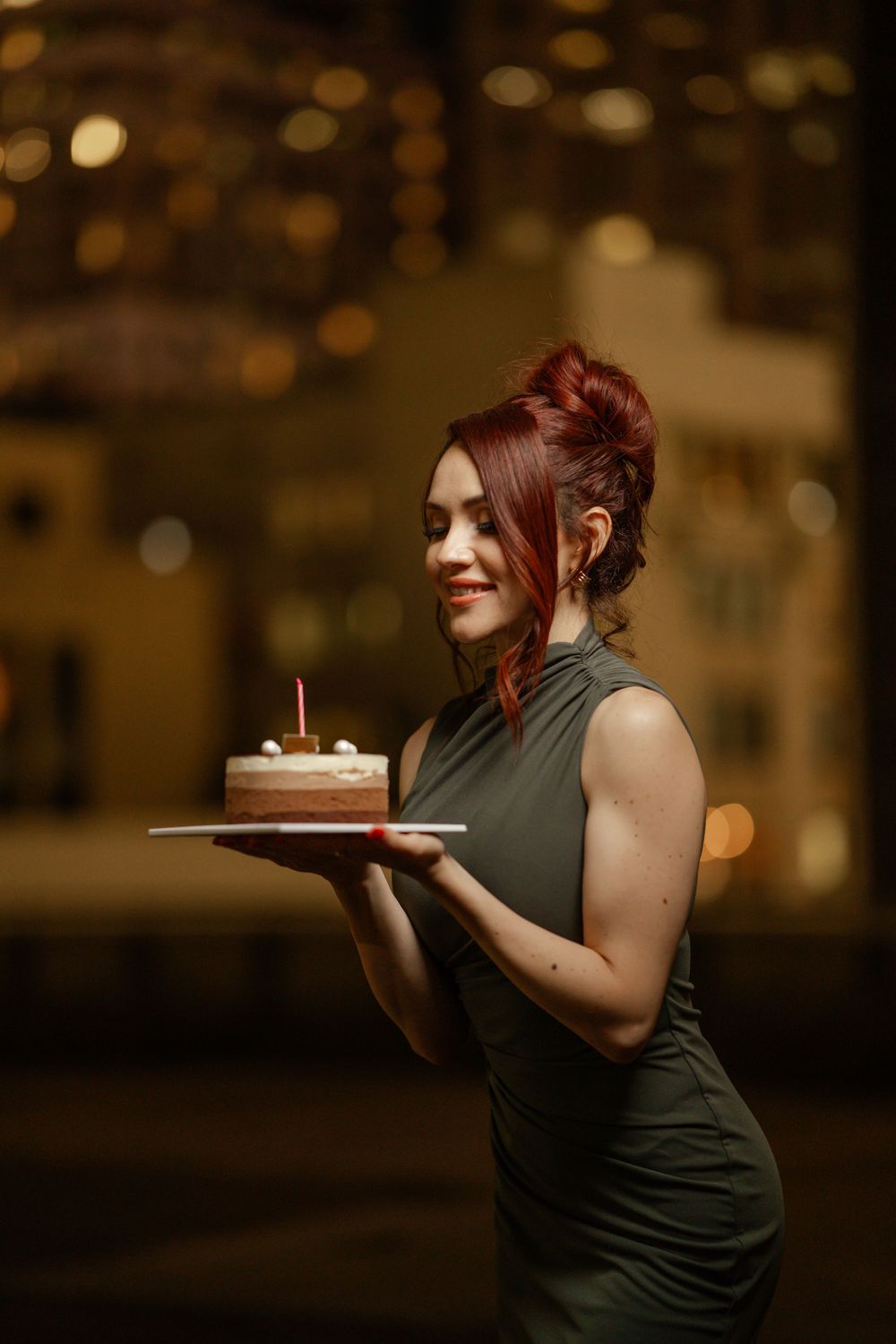 A woman in a dark evening dress stands holding a birthday cake with a candle against a blurred city backdrop.