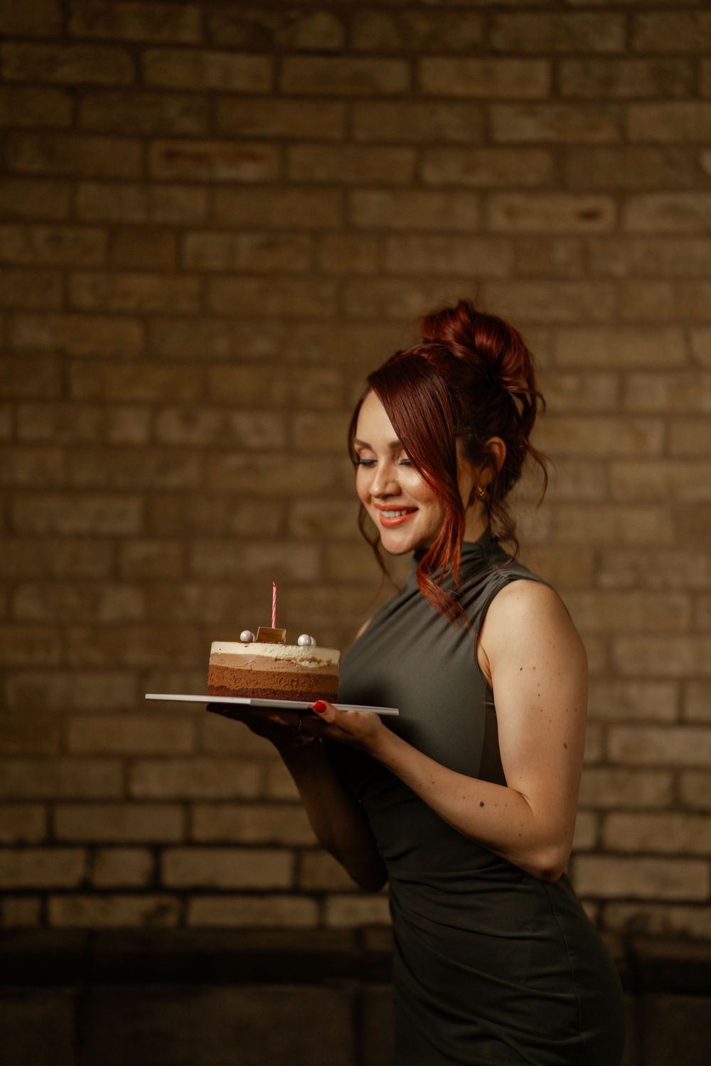 A figure in a grey sleeveless dress holds a birthday cake with candles against a brick wall background.