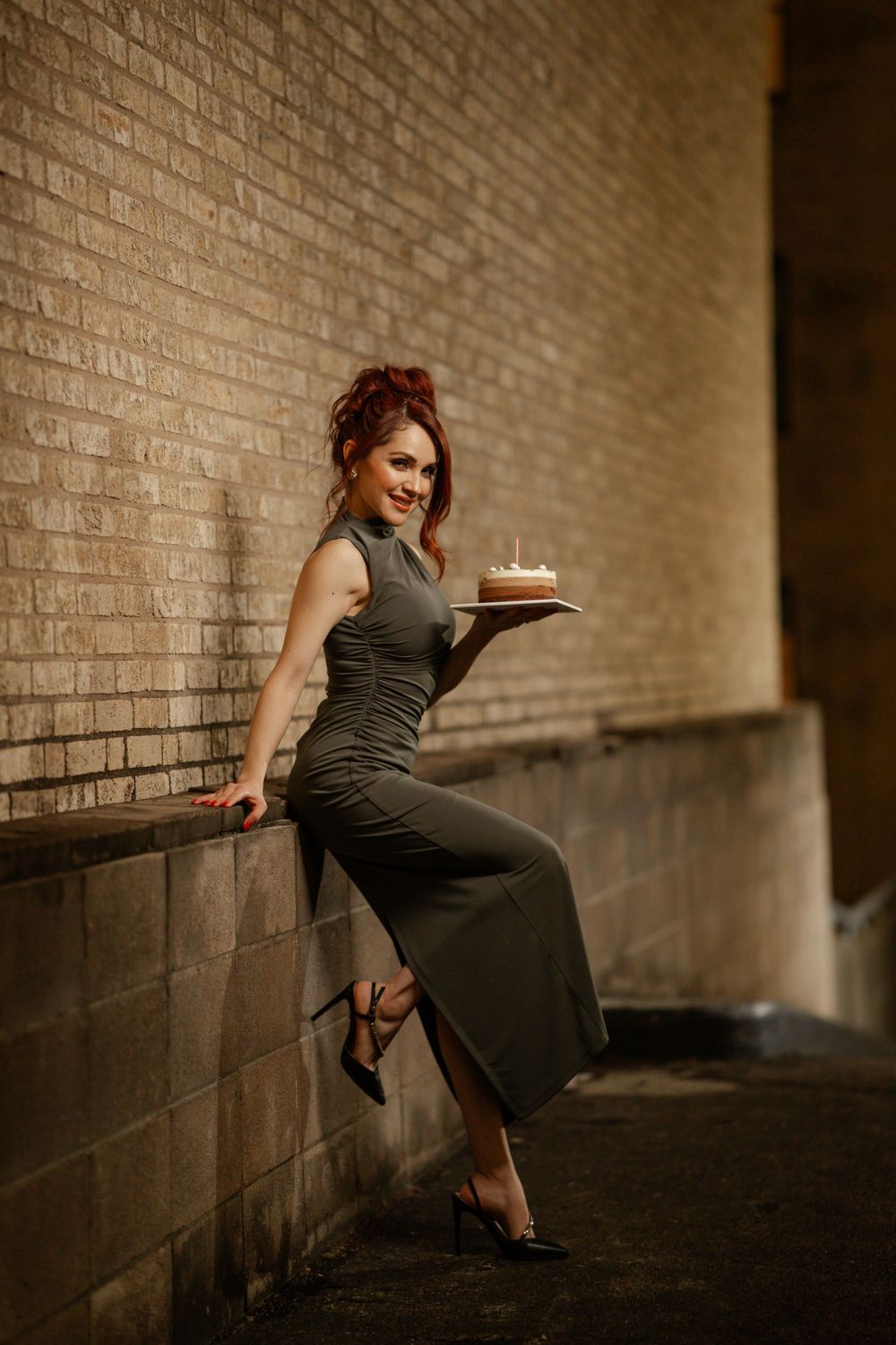 A woman in an olive green dress sits on a ledge against a brick wall holding a birthday cake with a candle.