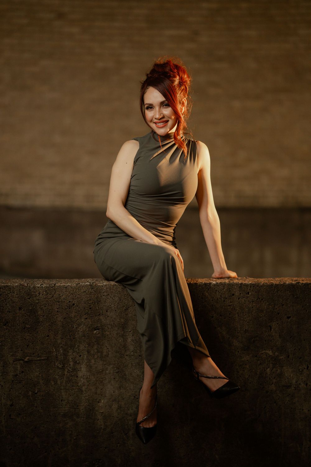 A woman in an olive green dress sits gracefully on a wall, illuminated by warm lighting against a dark background.