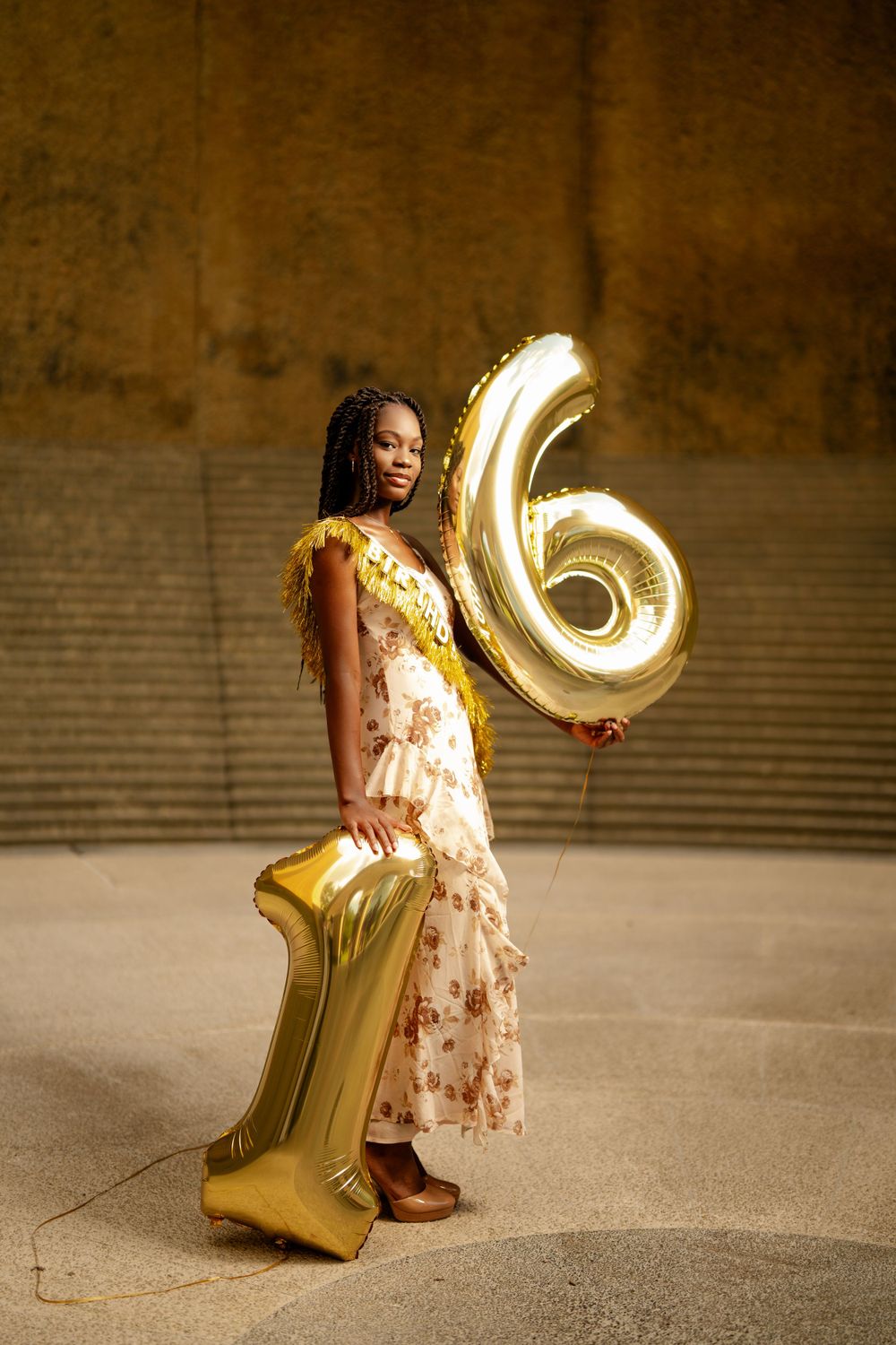 A person in a floral dress stands with large gold number balloons 1 and 6 against a brown textured wall.