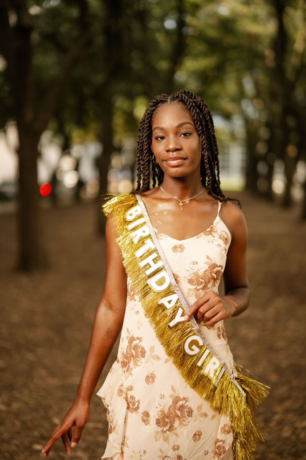 A woman in a cream floral dress and gold sash strikes a natural pose among trees in a park.