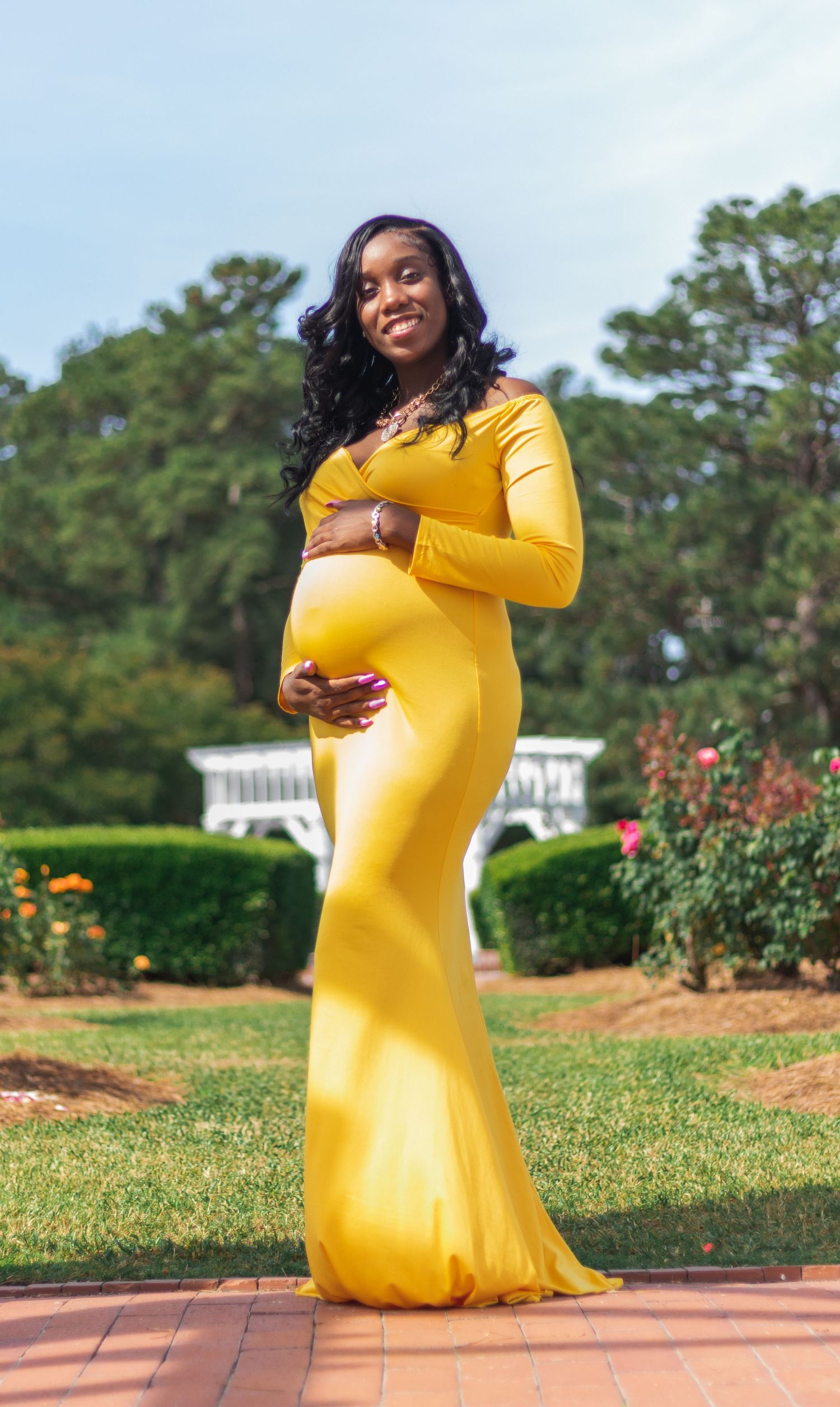 Expectant mother in long yellow dress poses gracefully in garden setting.