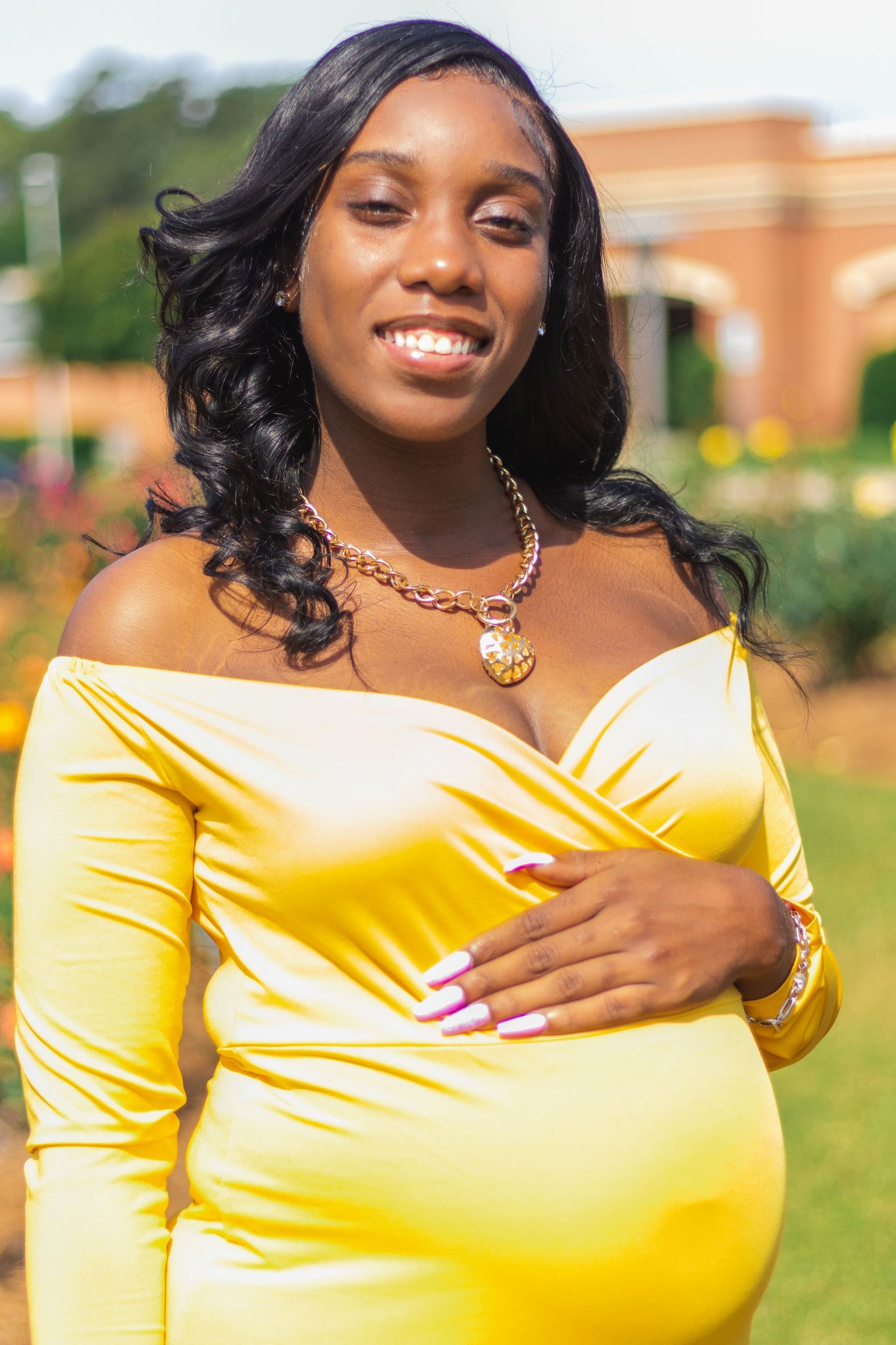 A glowing maternity portrait featuring a yellow off-shoulder dress and gold necklace against a garden background.