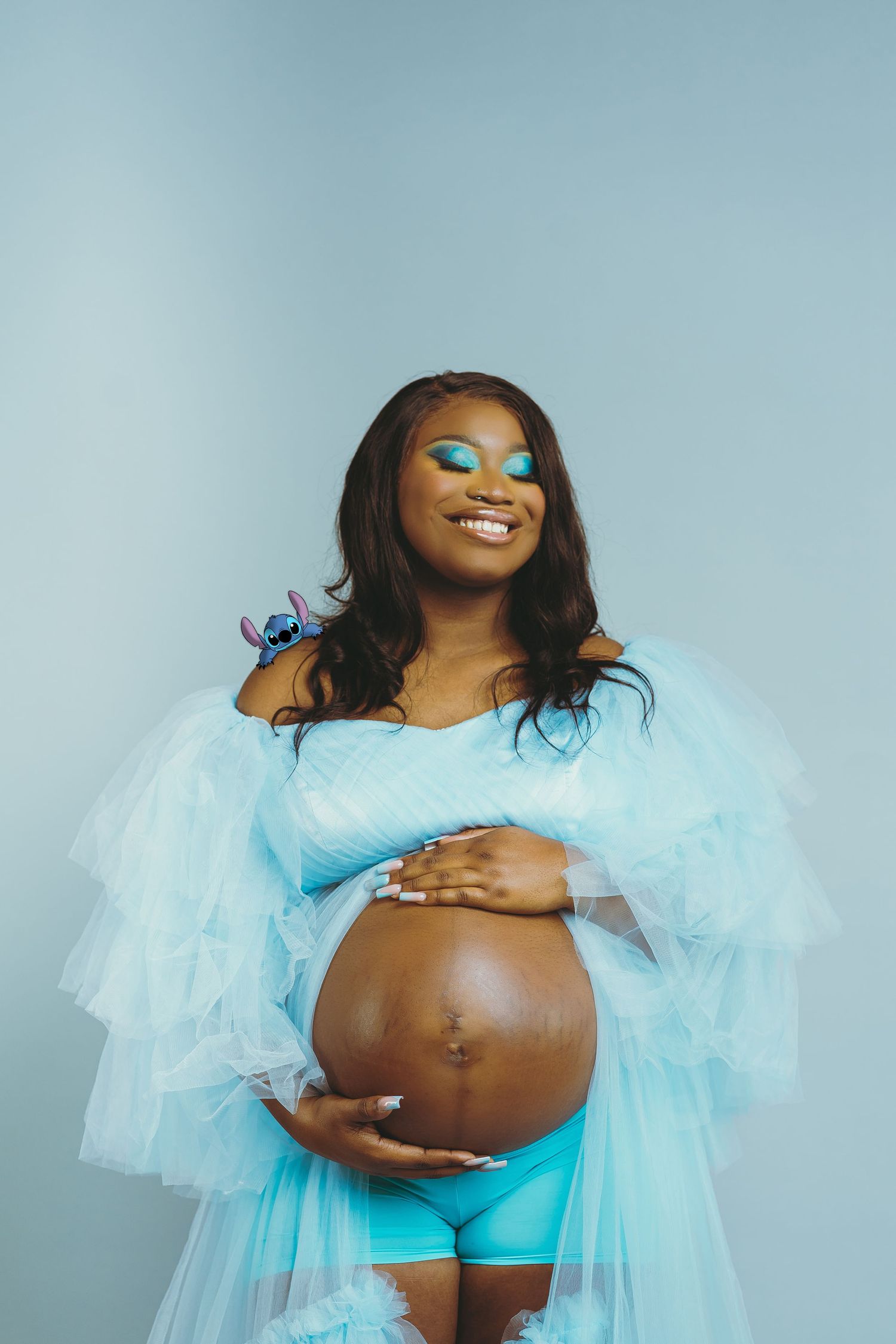 Maternity photo of woman in light blue tulle gown against blue backdrop.