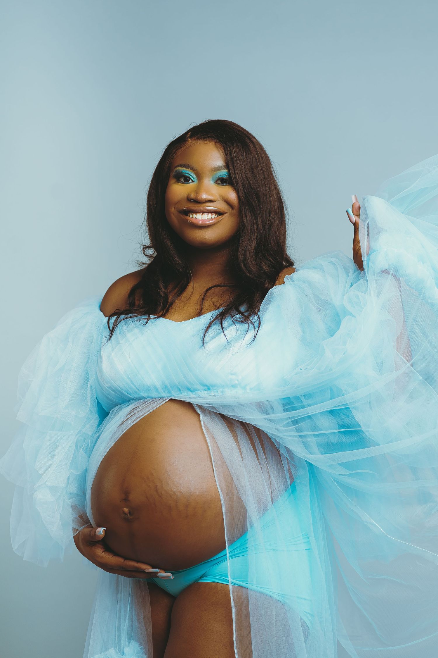 Expectant mother poses in flowing blue tulle dress against light background.