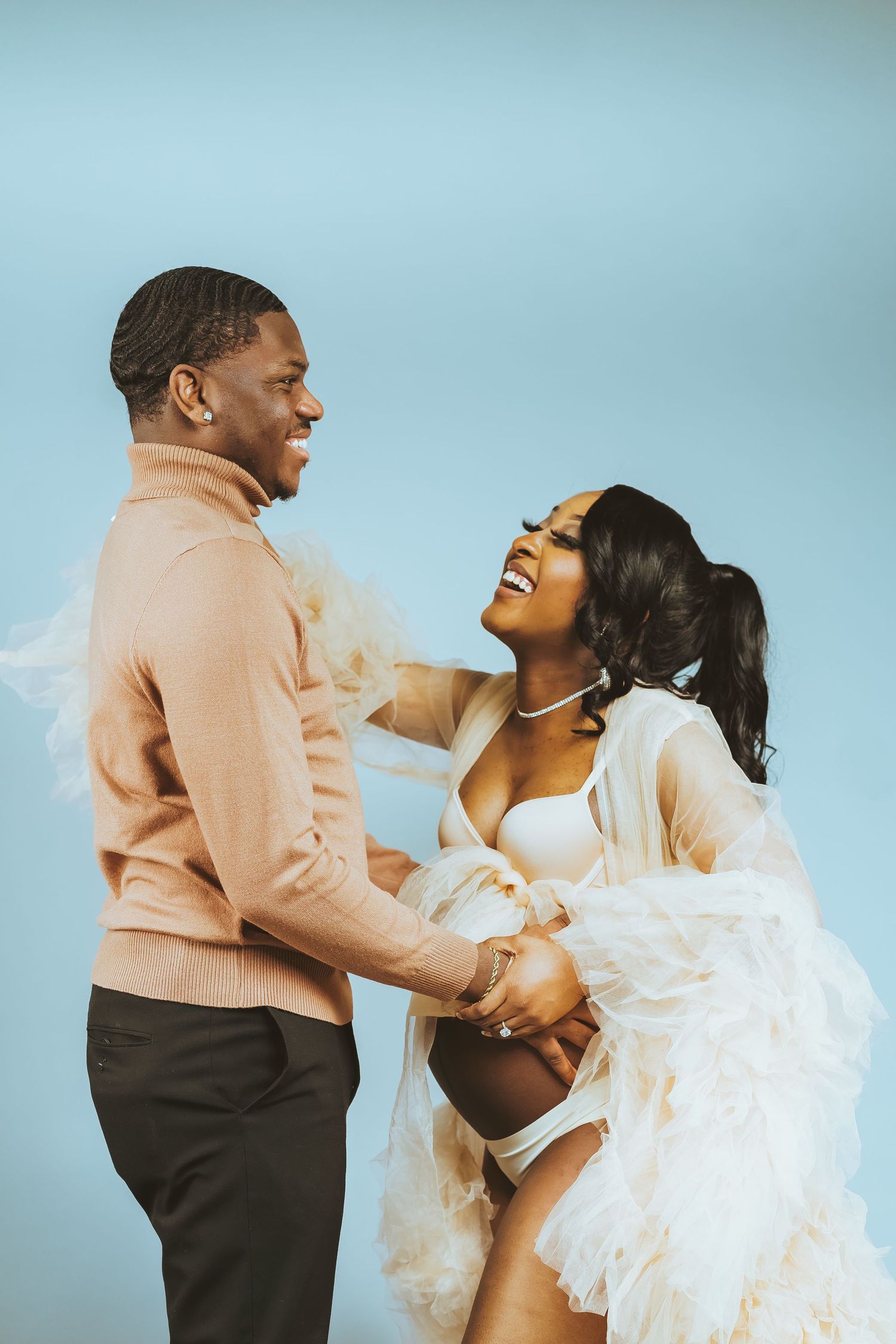 Couple shares intimate laughing moment during maternity photoshoot against blue background.