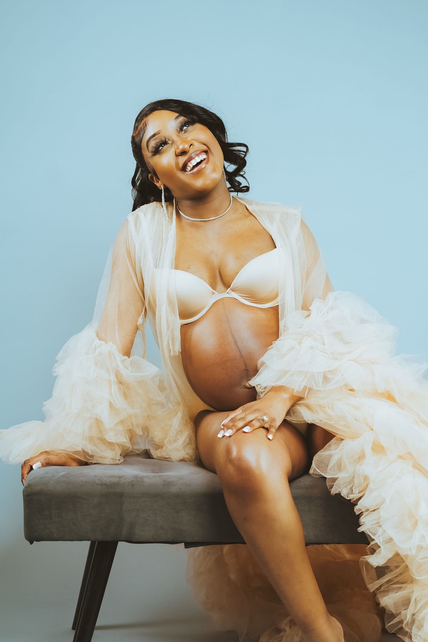 Maternity portrait featuring woman in white lingerie and flowing robe on bench.