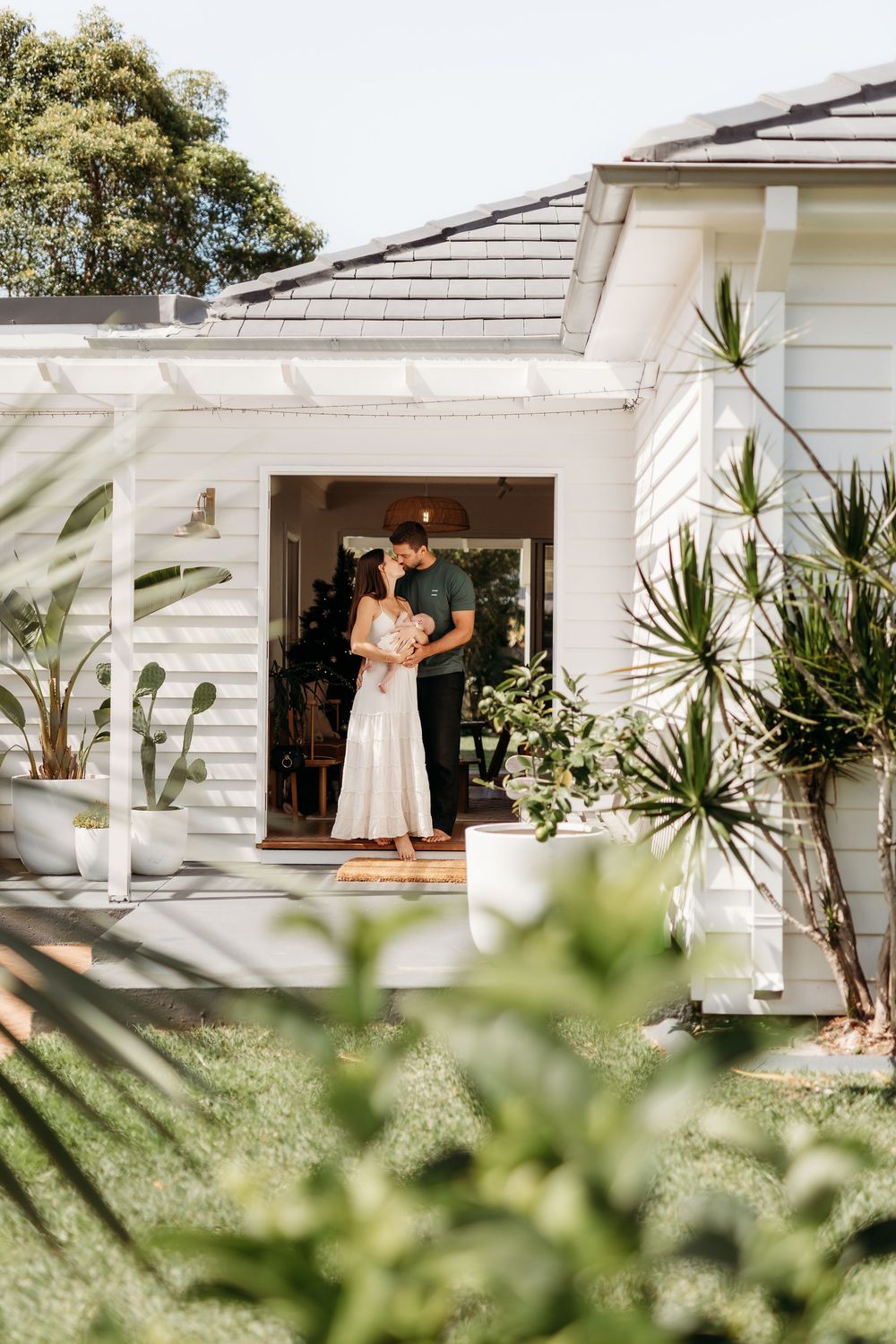 A family photographer taken at their front door at their home through the garden. The mum and dad are in love and cuddling their newborn.