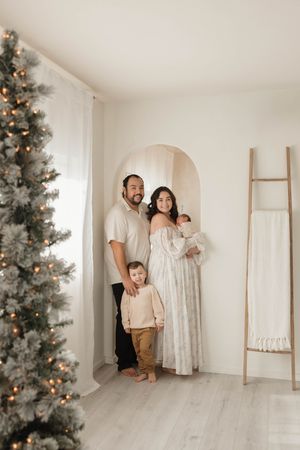 Family poses together in front of decorated Christmas tree in minimalist white room with wooden ladder decor.