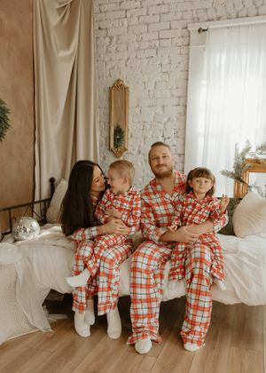 Family in coordinated red plaid sleepwear poses playfully on floor against white brick wall.